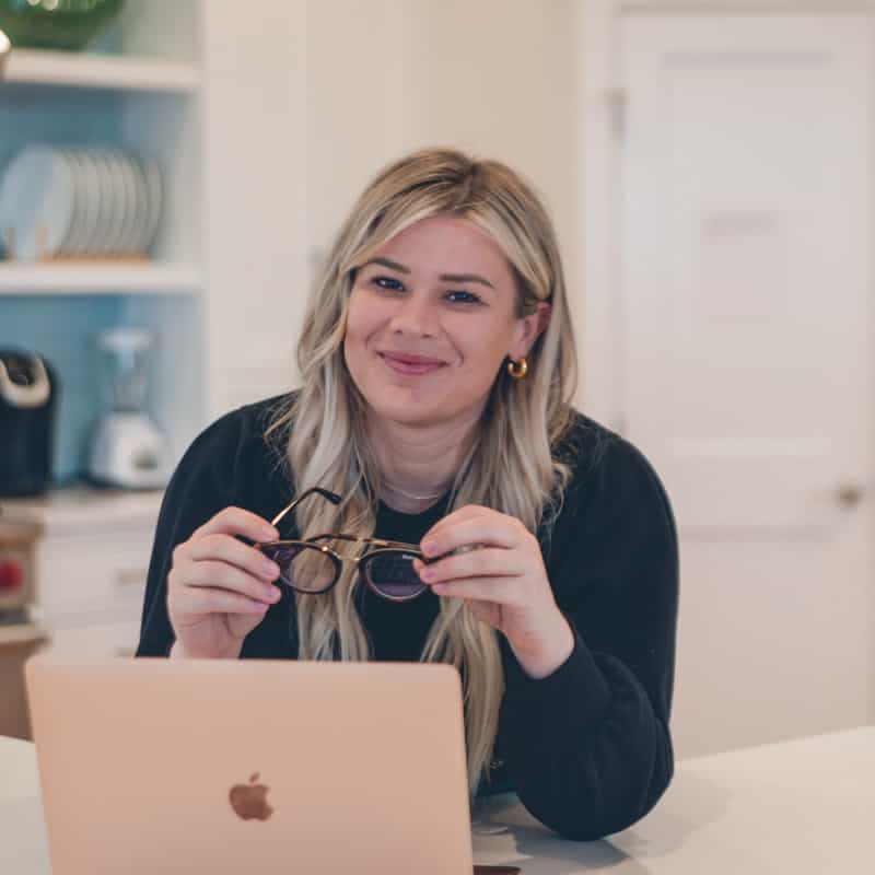 A person is smiling at the camera, holding glasses, with a laptop on the table in a home or office with a neutral-toned background.