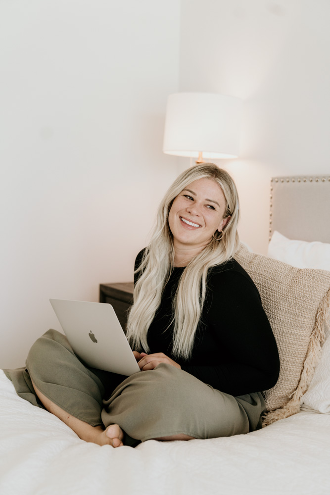 A person is smiling and sitting on a bed, holding a laptop. There's a lamp behind them, and the room has a cozy ambiance.