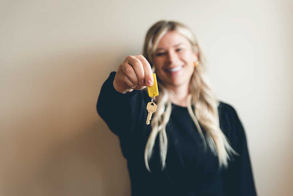 A smiling person holding out a set of keys with a yellow key fob, focus on keys, with a soft beige background.