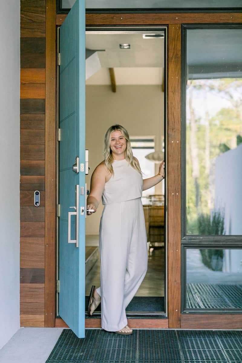 A person stands in a doorway, smiling, holding the door open, dressed in casual attire. A modern home interior and greenery are visible through the door.