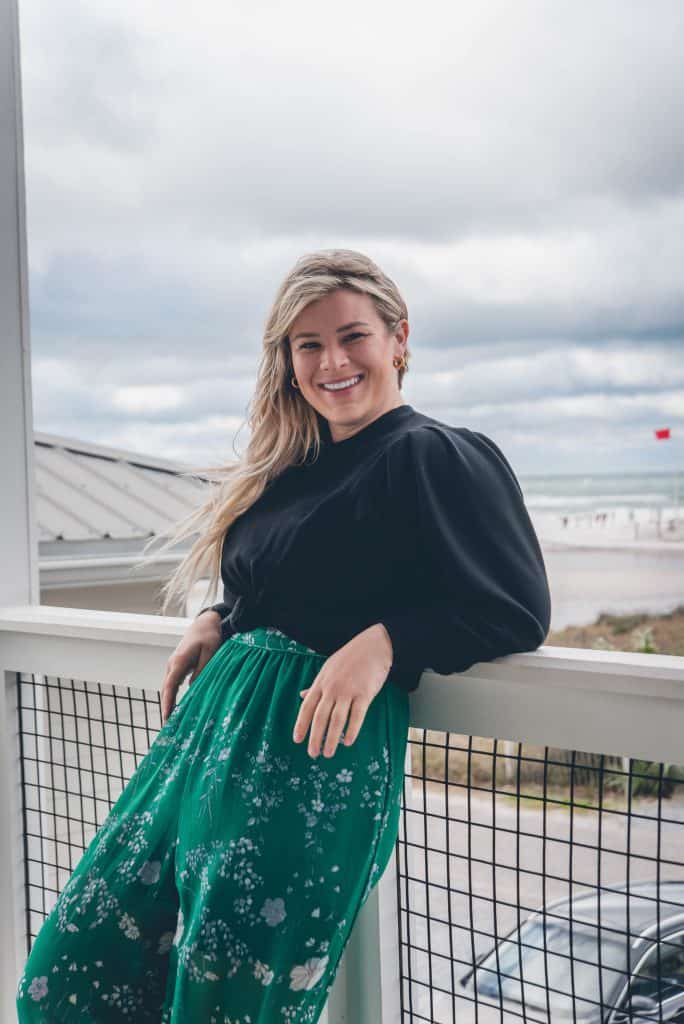 A smiling person leans against a balcony railing, wearing black top and floral green pants, with clouds and a coastal landscape in the background.