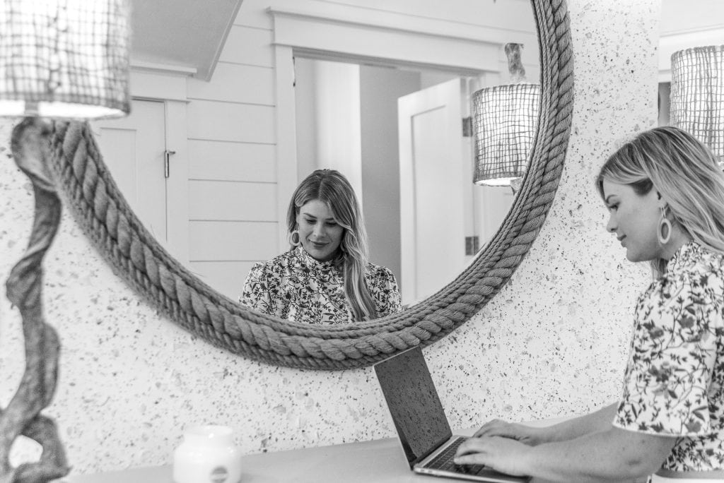 A person sits at a table working on a laptop, reflected in an oval mirror framed by a thick rope. It's a monochrome, well-lit room.