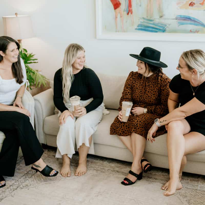 Four people are sitting on a couch, laughing and enjoying a conversation. They are holding drinks and dressed in casual attire, in a cozy room.