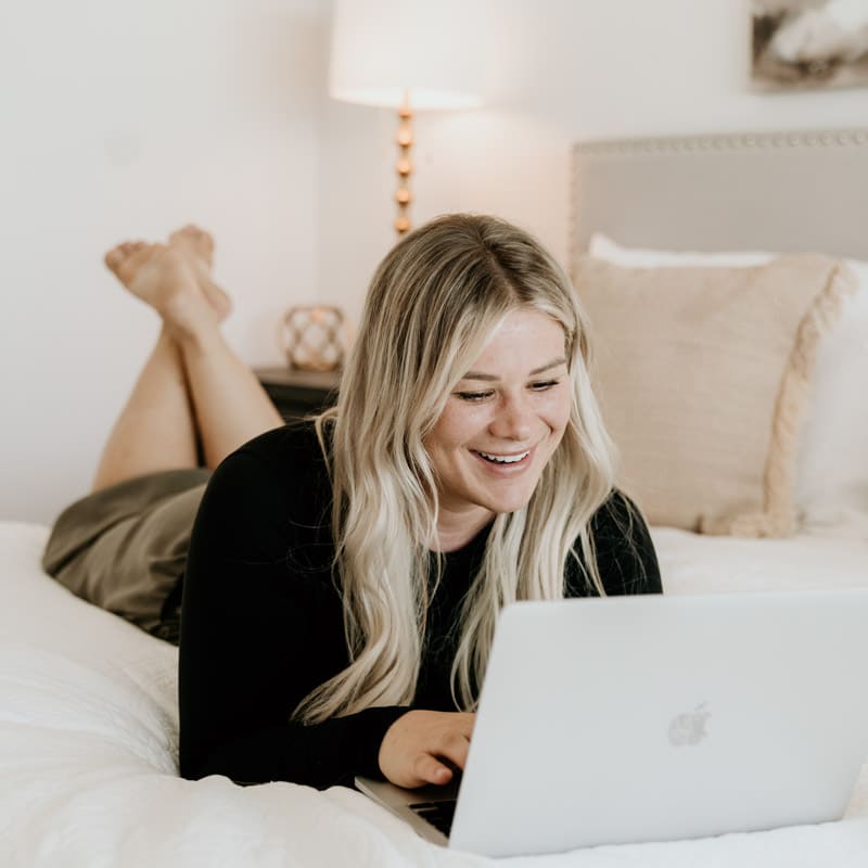 A person is lying on their stomach on a bed, smiling while looking at a laptop. The room has a warm, cozy decor.