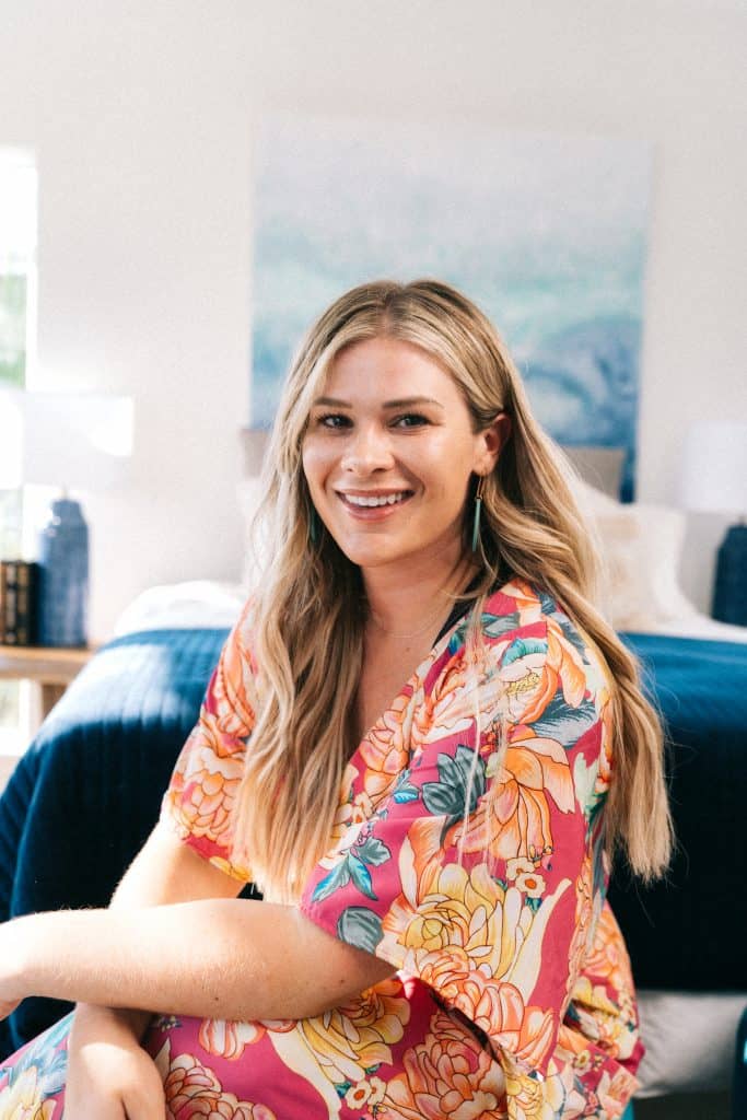 A person with blonde hair and a floral robe is smiling, seated indoors with a blue bed and abstract painting in the background.