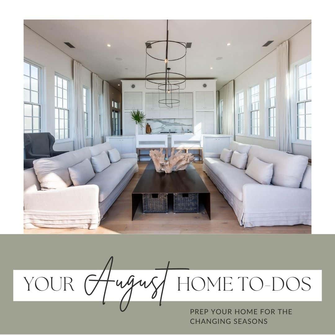 A modern, open-plan living space with large windows, neutral-toned sofas, a central wood table, a chandelier, and a kitchen in the background.