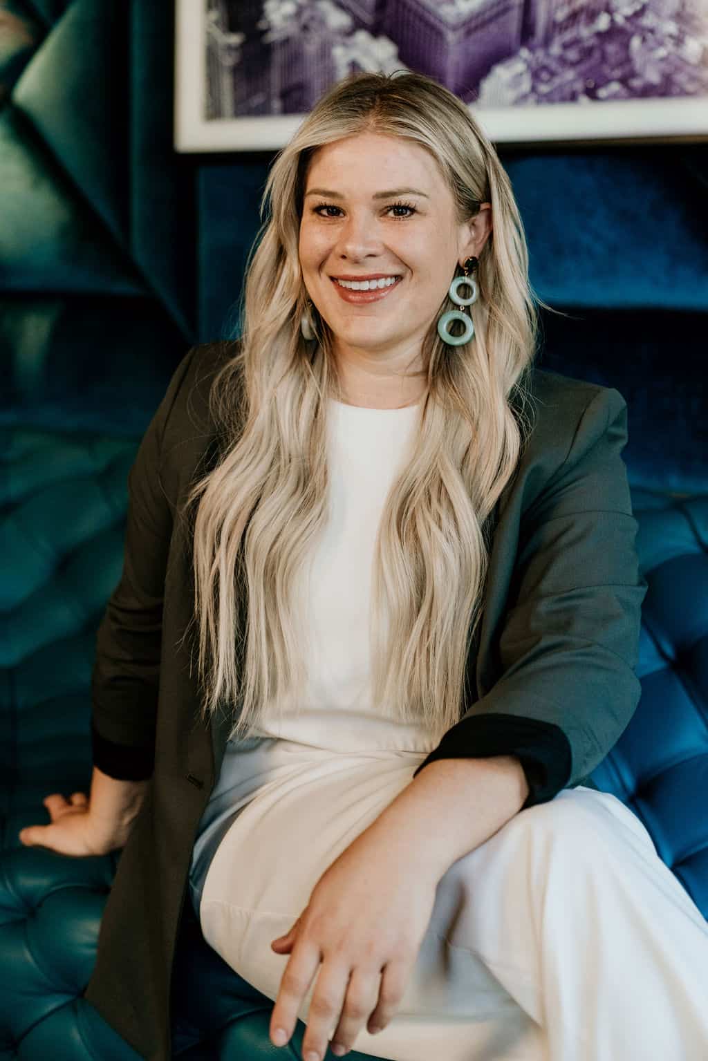 A smiling person in a green blazer and white outfit sits on a blue tufted couch with a vibrant purple floral picture in the background.