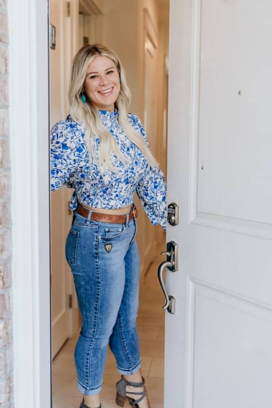 A person stands in a doorway, smiling, wearing a blue floral blouse, jeans, dangling earrings, and sandals, in a well-lit, neutral-colored hallway.