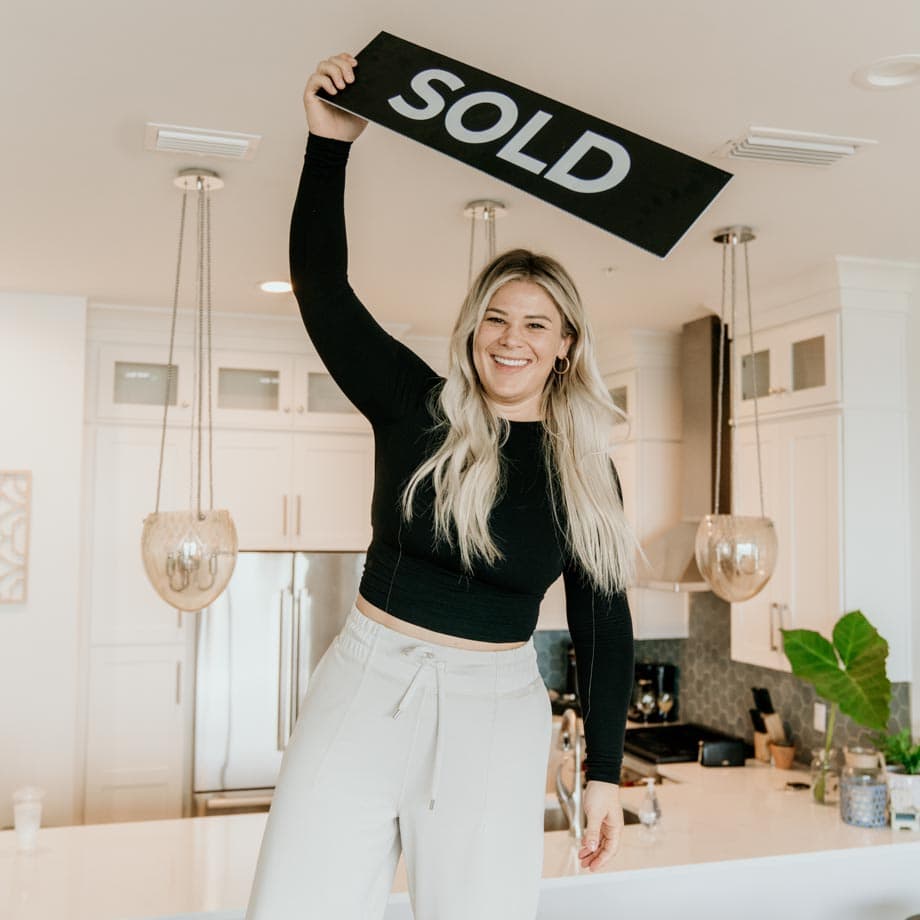 A person stands in a bright, modern kitchen, smiling and holding a "SOLD" sign overhead. They appear happy. Domestic setting with contemporary design.