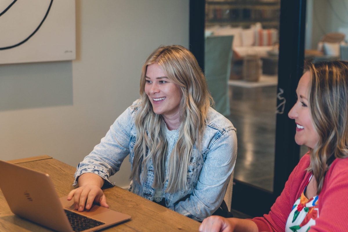 Two people are sitting at a wooden table, smiling and looking at a laptop screen. A modern interior and artwork are visible in the background.