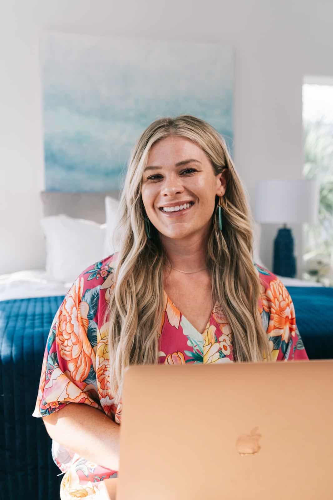 A smiling person with blonde hair, wearing a floral top, sits behind an open laptop. Abstract artwork and a blue bed adorn the background.