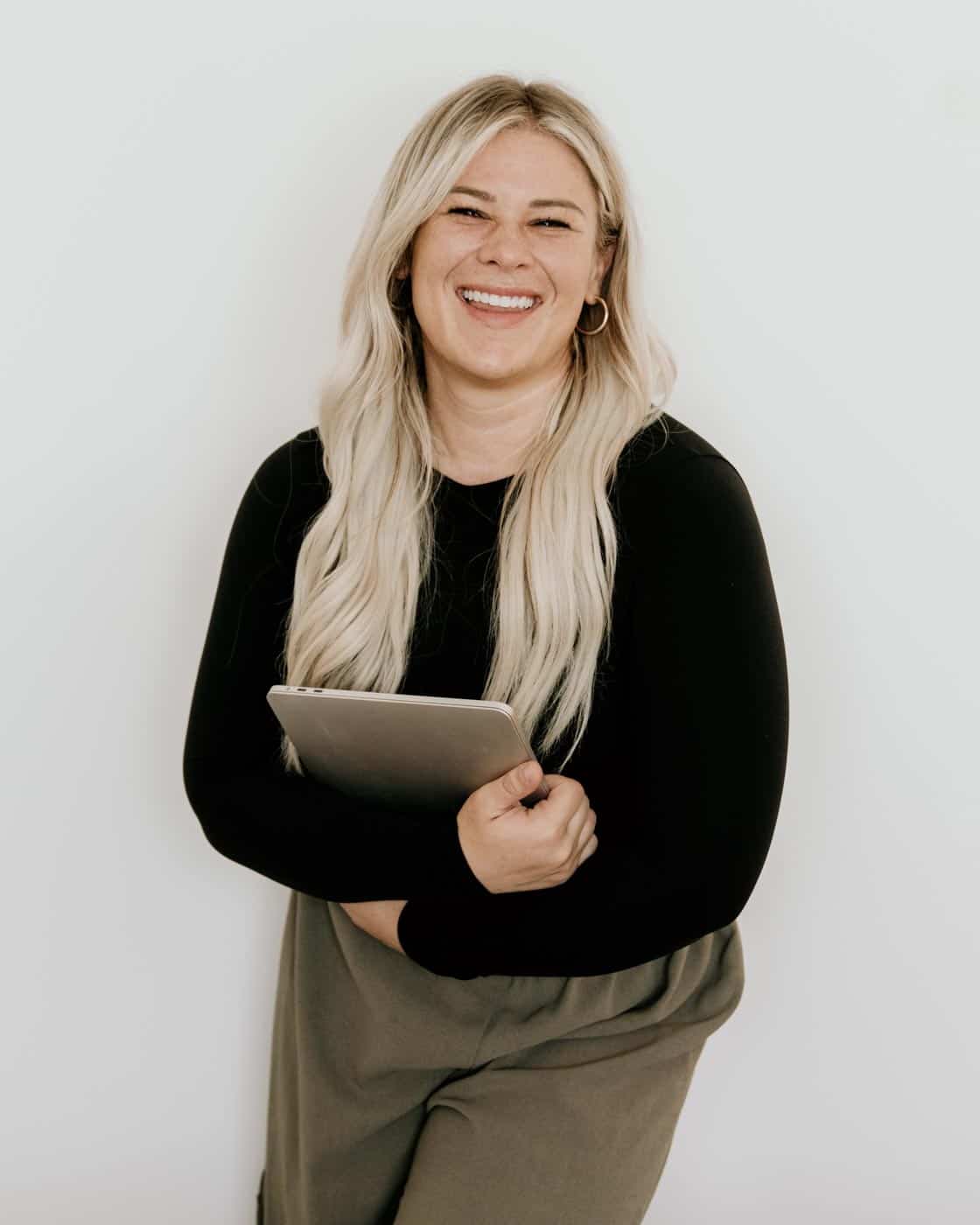 A smiling person with long blonde hair, wearing a black top and olive pants, holds a tablet, standing against a white backdrop. They exude a friendly demeanor.