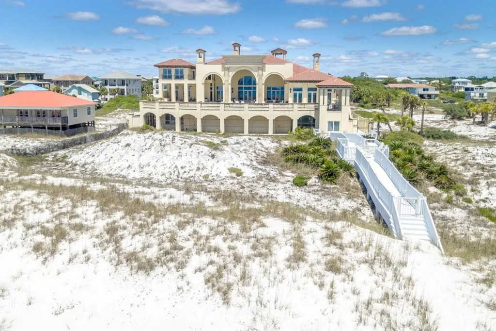 A large, luxurious coastal house with multiple balconies overlooks a sandy beach, connected by a wooden walkway, flanked by smaller houses and vegetation.