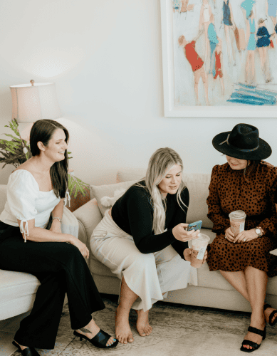 Three people sit on a couch, laughing and enjoying coffee, with stylish clothes and a bright painting in the background. The ambiance is cozy and fashionable.